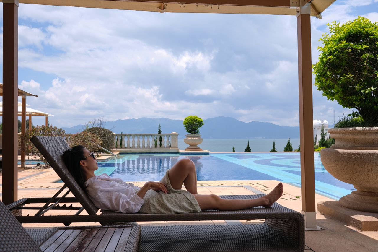Mastering the First Impression: Your intriguing post title goes here A woman in sunglasses relaxes on a sunlounger by a luxury poolside with a scenic view.